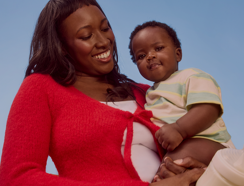 Woman in a red sweater holding a baby and a formula bottle against a clear blue sky